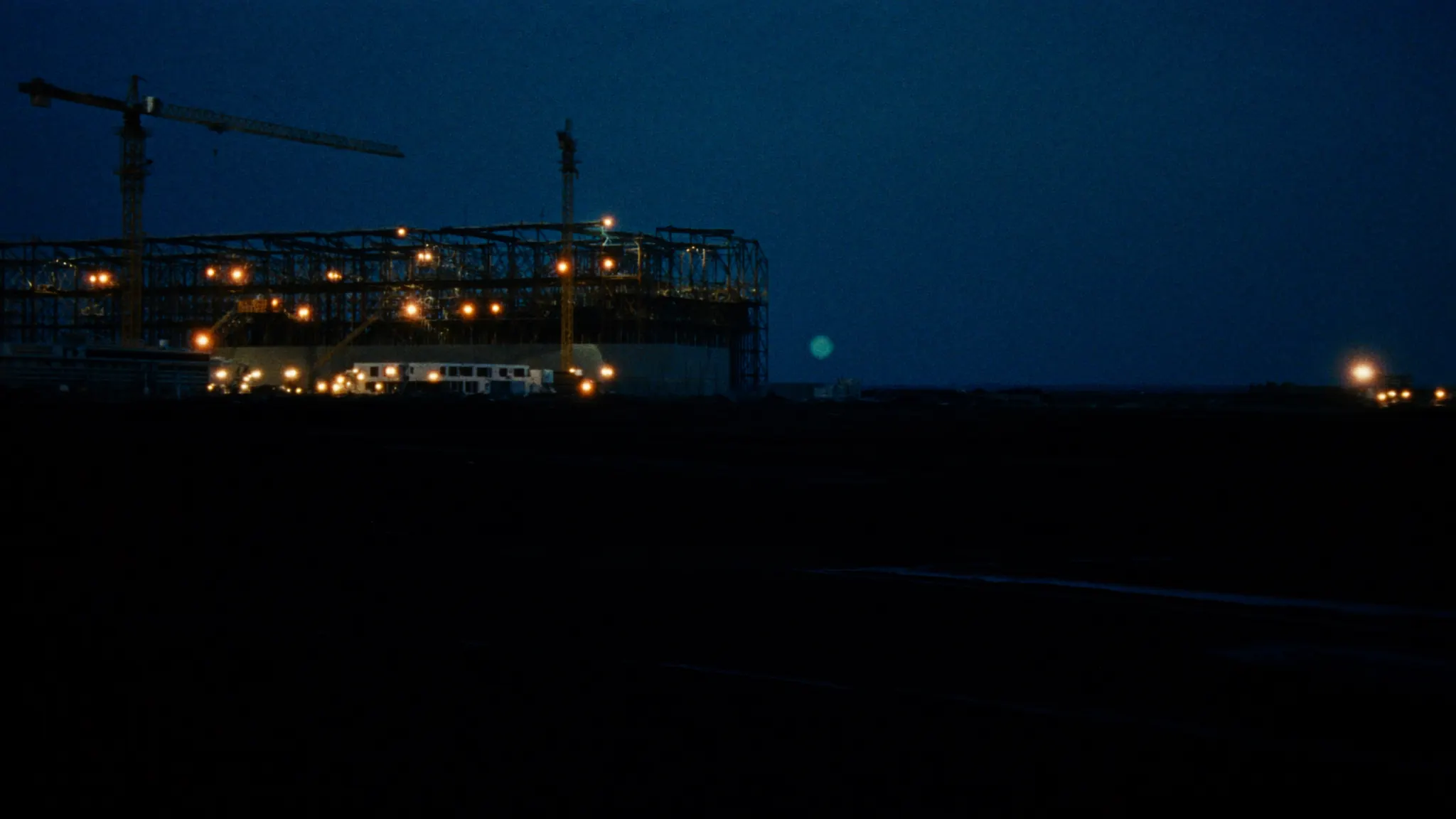 A large electrical construction site at blue hour, cranes and steel structure lit by scattered work lights across open ground.
