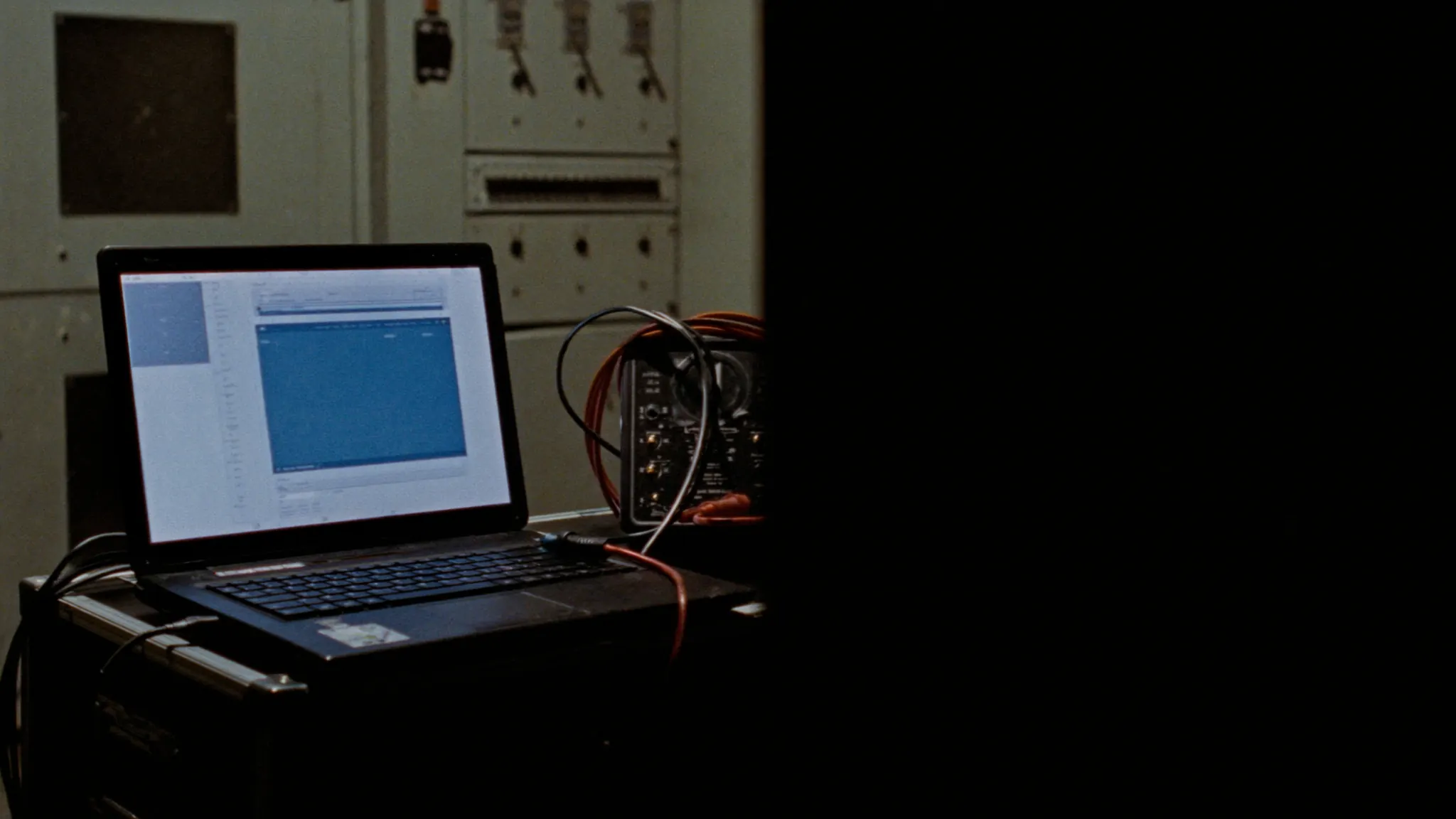 A rugged laptop open on an equipment case beside a portable insulation test set, screen glowing in a dark switchgear room.